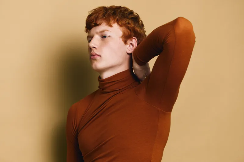 Young man with red hair wearing brown turtleneck posing with hand behind head against beige background. Fashion, portrait, style and beauty with casual outfit in studio lighting.