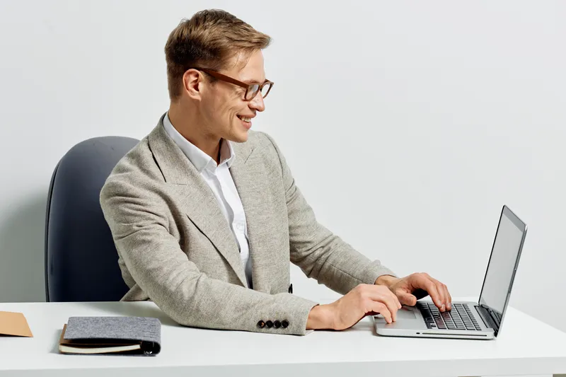 Smiling young man with glasses working on silver laptop at white desk in modern office environment, dressed in light gray blazer and white shirt, casual business style, positive mood, technology conce