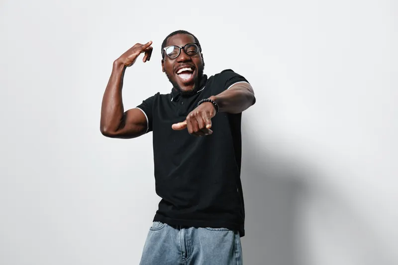 African American man with dark skin wearing glasses and black polo shirt laughing and pointing while holding a cookie in studio with white background.