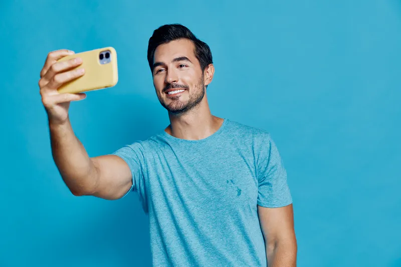 Smiling man taking a selfie with mobile phone, wearing a blue t-shirt, against a bright blue background, showcasing a fun and casual vibe.