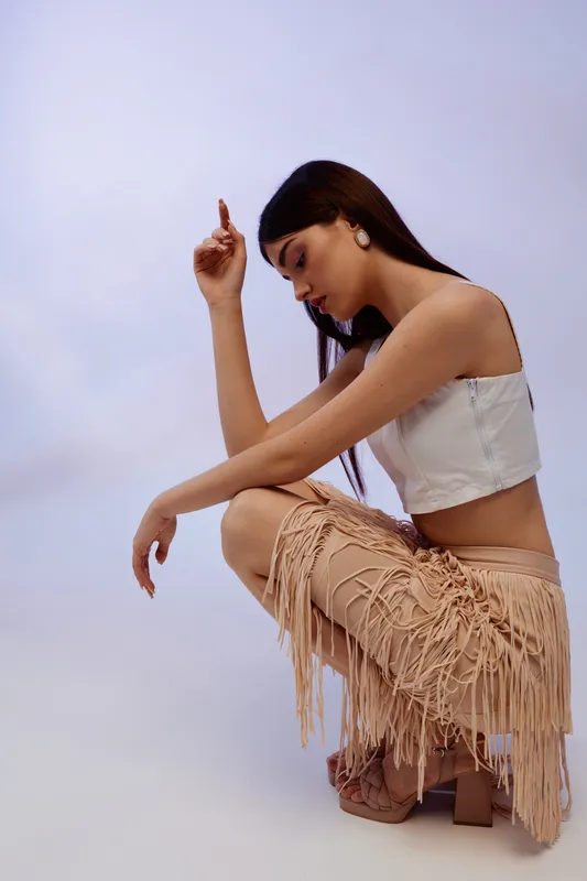 Young woman in fashionable beige fringe skirt and white crop top poses in studio with soft lighting. Concept of beauty, fashion, and modern style for editorial or advertising.