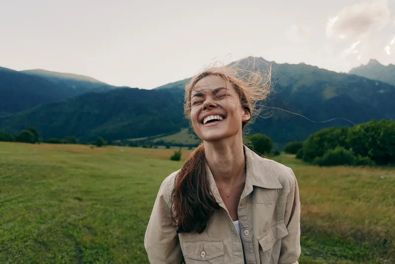 Young woman with curly hair smiles broadly in a sunny field with distant mountains, conveying a carefree outdoor mood and a sense of warmth and joy.