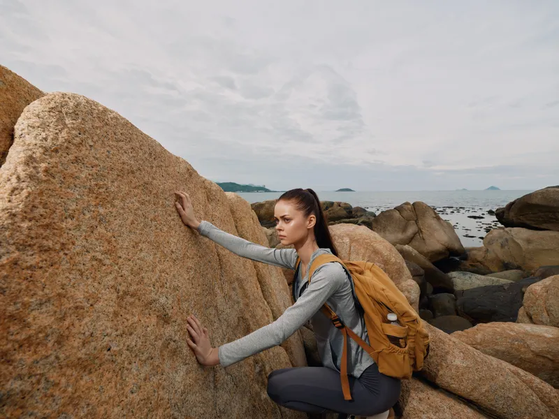 Young woman climbing rocks by the seaside, wearing casual outdoor attire with a backpack, focused on adventure in a natural landscape