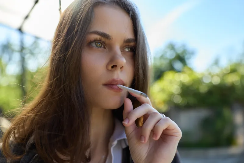 Woman outdoors smoking a cigarette, casual lifestyle portrait with thoughtful expression, natural sunlight, blurred green background, conveys calm reflective mood outdoors