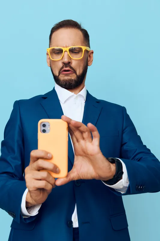 A confident business professional man takes a selfie on his phone, posed against a solid color backdrop, conveying focus, curiosity, and assertive emotion in a modern work scene.