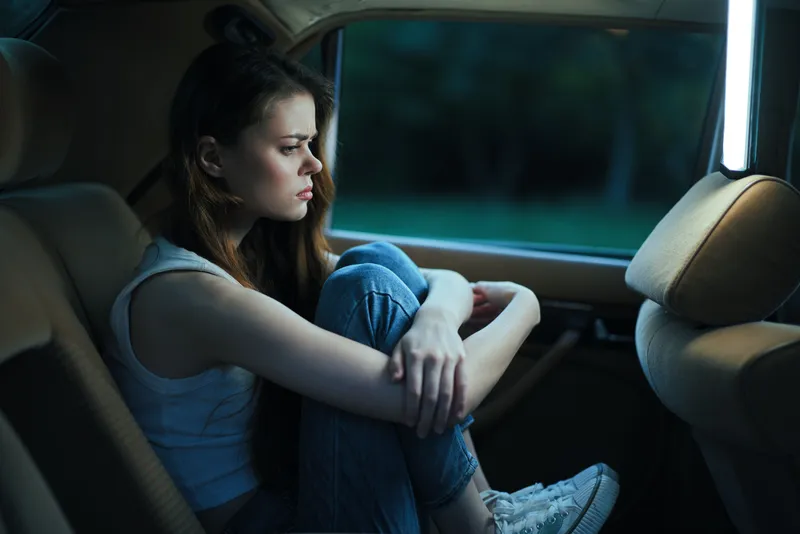 A young woman with long hair, wearing a sleeveless top and jeans, sitting pensively in the backseat of a car with a somber expression, reflecting emotions of solitude and introspection.