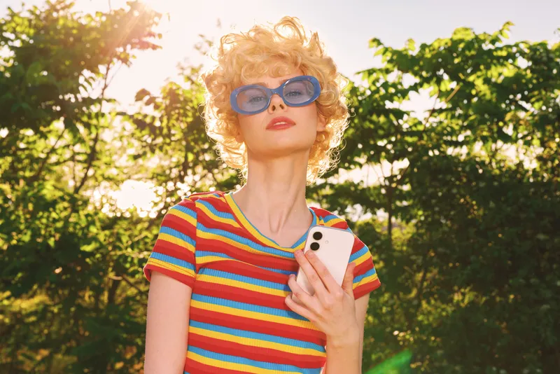 Smiling woman with short curly hair wearing a colorful striped shirt and blue sunglasses, holding a smartphone outdoors in bright sunlight, retro film color, lifestyle portrait.