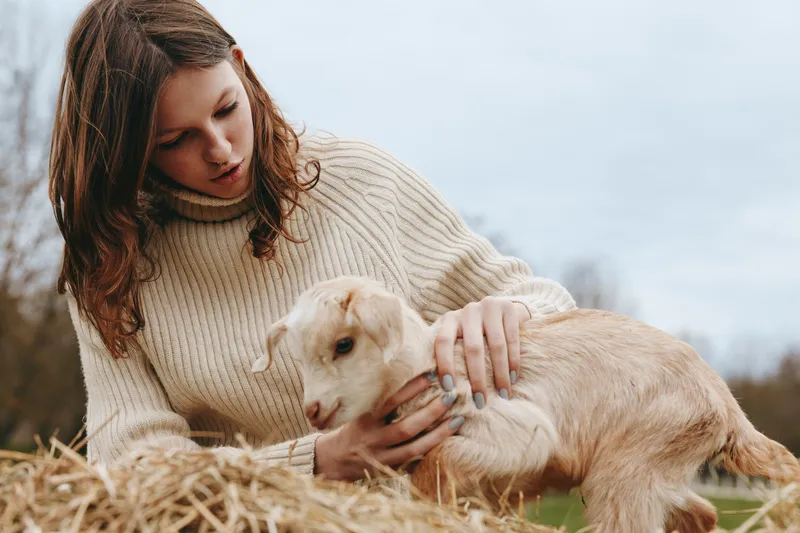 Goat kid lifestyle outdoors woman pet farm rustic gentle connection, a candid outdoor scene showing a woman with a goat kid among hay, soft light, calm mood, natural colors, joyful companionship