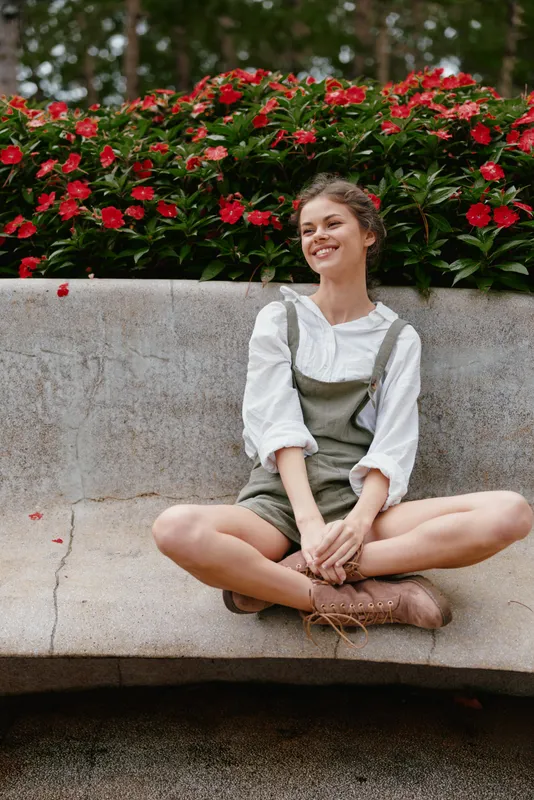 Young woman in casual olive green overalls sitting in a garden surrounded by blooming flowers, exuding joy and tranquility in a vibrant, natural setting