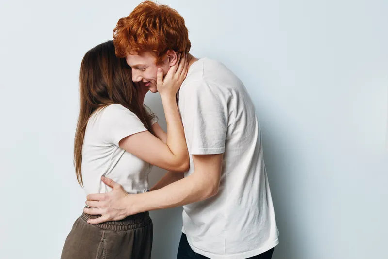 Couple, love, affection and relationship with young woman and man embracing, smiling and touching faces on white background in casual clothing at studio.