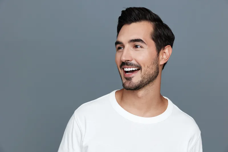 Cheerful smiling young man with stylish haircut wearing white t-shirt looking sideways, radiating positivity and confidence against a plain gray background.