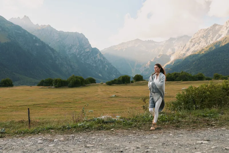 A woman walks along a gravel path in a mountain valley, wearing a light shawl, enjoying outdoor lifestyle amid serene nature and expansive scenery, capturing a calm travel moment
