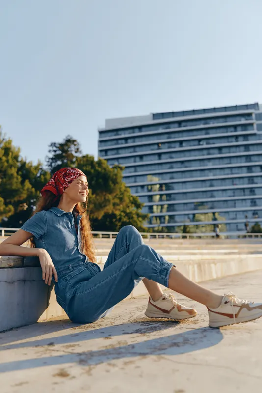 Relaxed woman wearing denim jumpsuit and red headscarf sitting outdoors in urban environment, enjoying sunny day with modern building and greenery background.