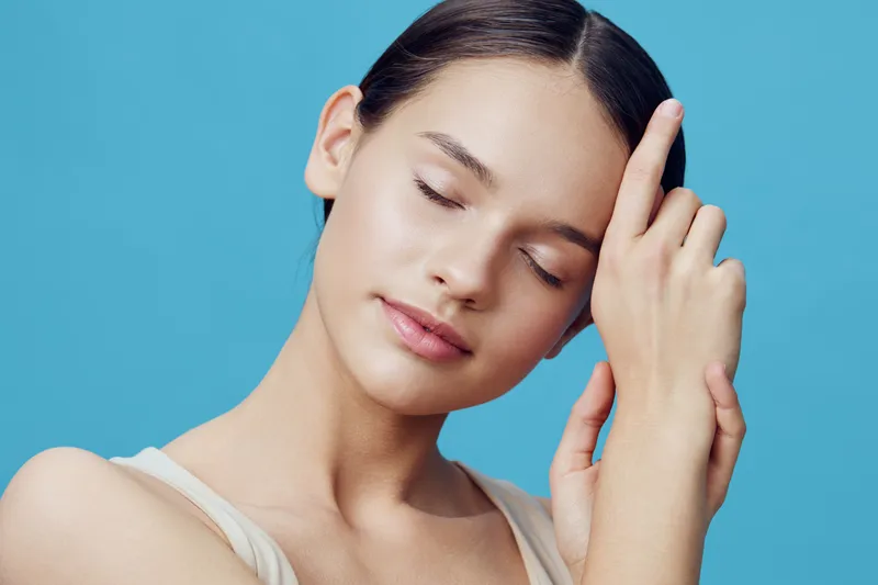 Serene young woman with closed eyes and soft smile against a blue backdrop, embodying calmness and tranquility with a natural beauty style