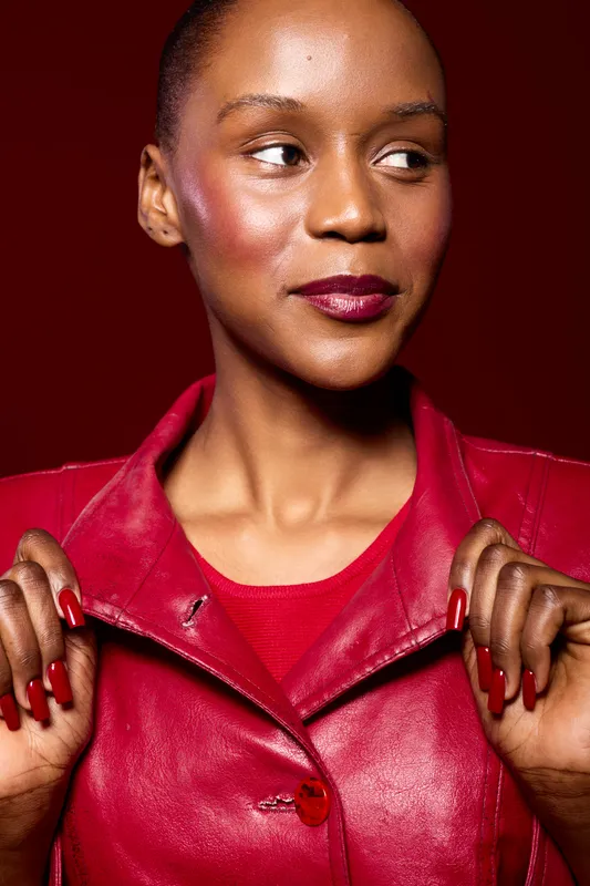 Confident dark skinned African woman in red leather jacket looks aside with slight smile. Studio portrait on red background. Concept of fashion, beauty, style, and female empowerment.