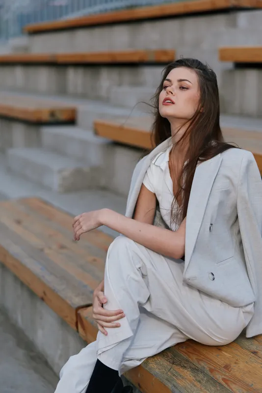 Young woman in light suit sitting on wooden steps looking confident and contemplative in urban outdoor setting with neutral tones and soft natural light