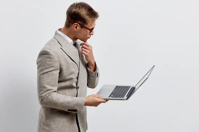 Businessman wearing glasses in a gray checked blazer holding a silver laptop while thinking, isolated on a plain white background. Professional work concept, modern technology, office environment, ser