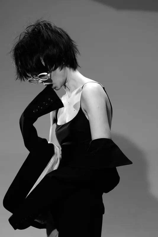 Black and white portrait of a young woman with short hair wearing glasses and a stylish coat, exuding chic fashion and elegance.