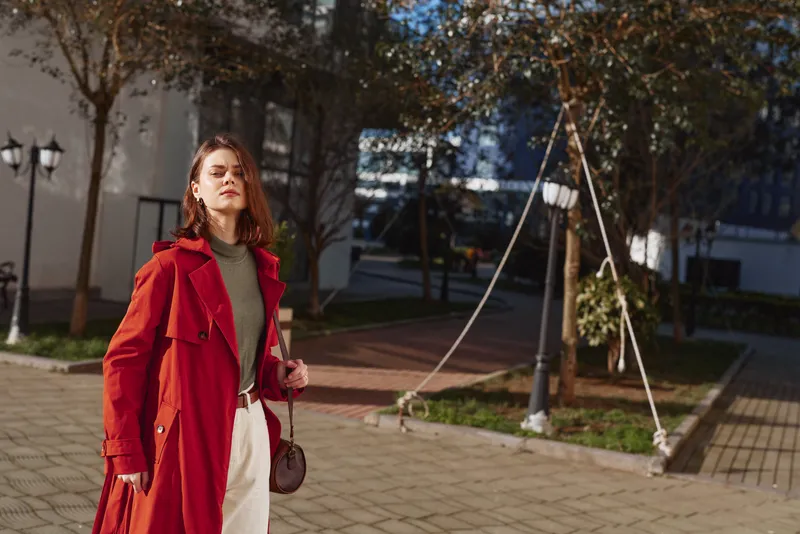 Young woman in a red coat stands confidently outdoors on a sunny day with eyes closed enjoying the warmth and fresh air in an urban park setting.