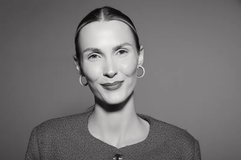 Portrait of a smiling Caucasian woman with light skin and slicked back hair wearing earrings and a textured top. Studio lighting on grey background.
