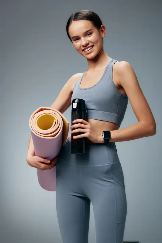 Fitness woman in athletic wear holding a yoga mat and water bottle, ready for training. Clean studio background highlights dedication, balance, and a healthy lifestyle during sport practice.