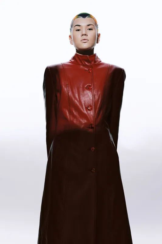 Woman with short hair wearing a long red leather coat standing confidently against a bright white background with sharp minimalist lighting.