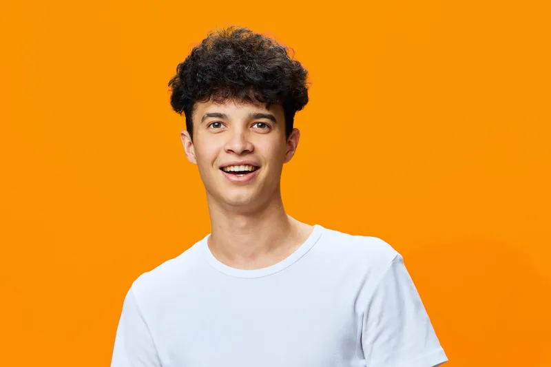 young man with curly hair smiling against bright orange background, wearing plain white t-shirt, cheerful expression, casual style for modern portrait.