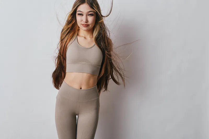Young woman with long hair wearing beige seamless fitness outfit in studio. Female athlete posing with natural makeup and casual hairstyle on neutral background for sport or active lifestyle.