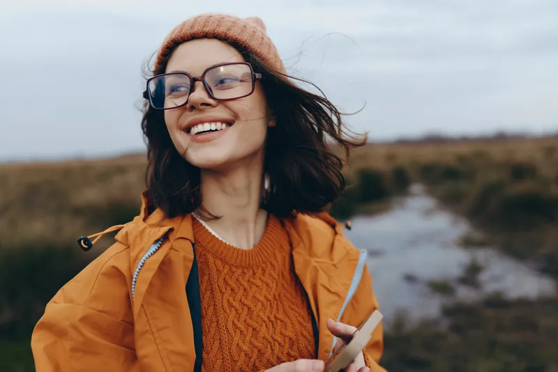 Woman smiling in a beanie and glasses, wearing a jacket, holding a smartphone outdoors by a river, casual lifestyle, soft daylight, calm nature, travel mood, gentle wind, autumn colors