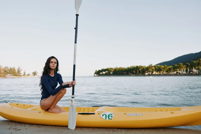 Female kayaker sits on a bright yellow kayak on a quiet sandy beach, holding a paddle upright as calm sea and distant hills create a tranquil coastal scene.