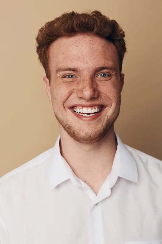 Smiling young man with red hair and freckles wears white shirt against beige background. Close-up portrait shows happy expression and natural skin texture. Concept of joy and confidence.