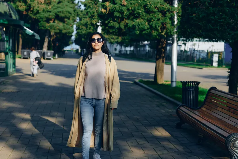 Woman walking outdoors in a long coat and sunglasses along a sunlit park street, casual street fashion, relaxed mood, daytime urban scenery, stylish yet comfortable look on a calm city day