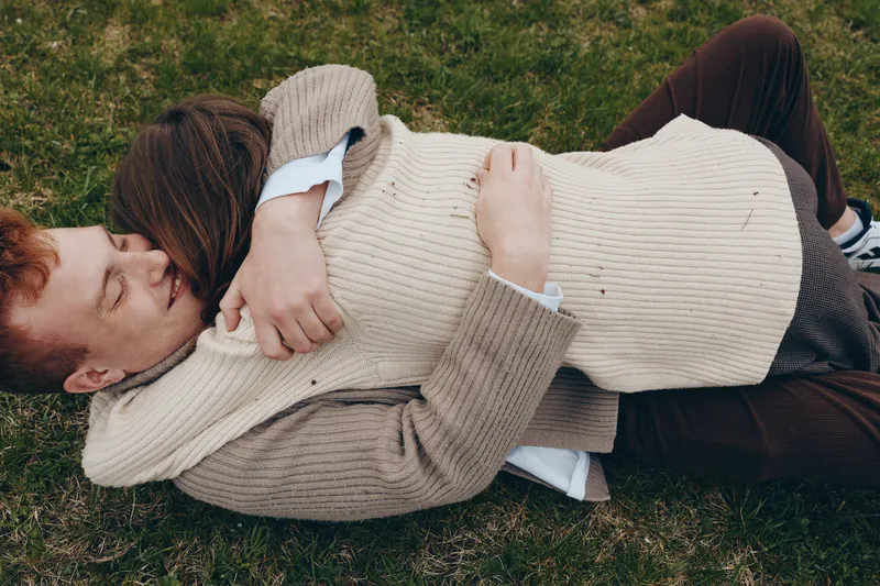 Couple lies on grass, wrapped in cream sweaters, sharing a quiet cuddle in a sunlit park. Cozy, intimate mood emphasizes warmth, affection, and relaxed togetherness.