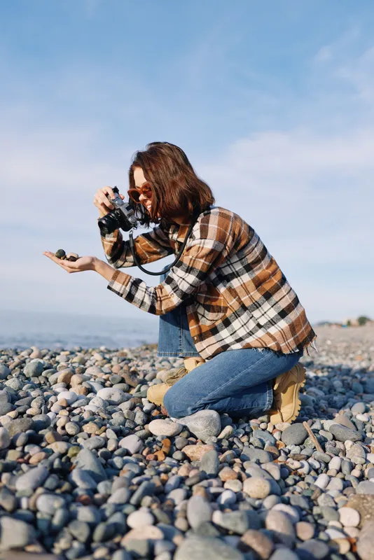 Photographer kneels on a rocky beach, aiming a camera with care and focus to capture candid coastal scenery, light breeze, and authentic moments in natural daylight.