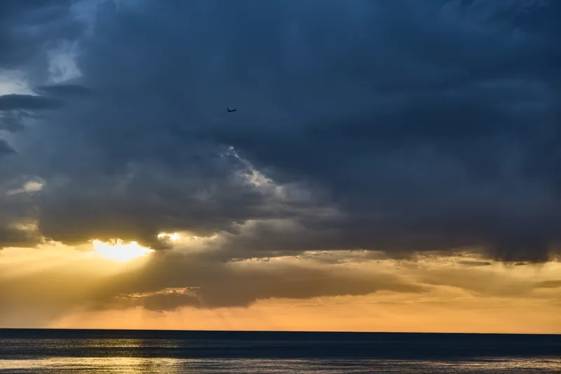 Dramatic sunset over ocean with dark storm clouds and golden sunlight breaking through, capturing nature's contrast between calm sea and turbulent sky