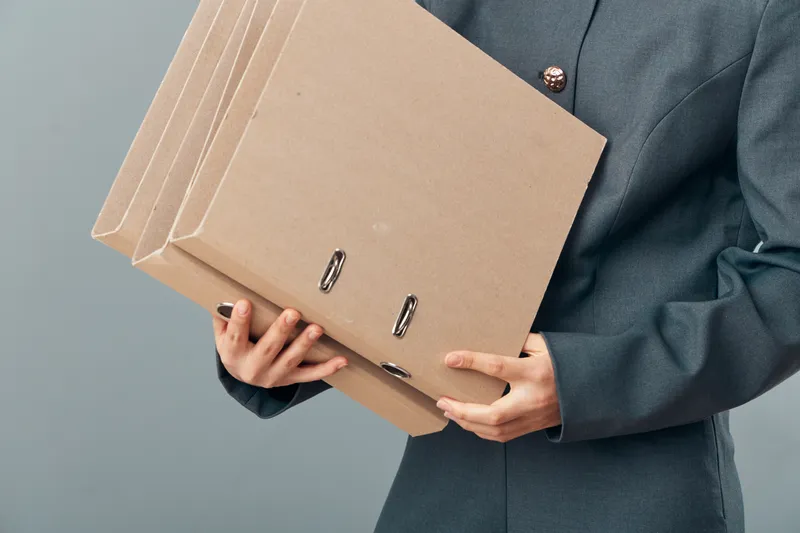 Person in formal suit carrying multiple cardboard folders for office organization, documents storage and paperwork management in professional environment with neutral background.