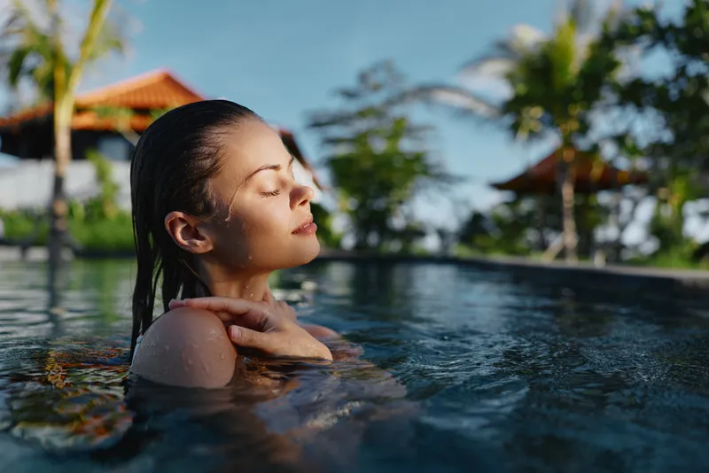Relaxed woman in a sunlit pool outdoors, eyes closed, serene expression, warm breeze, clear water, tropical greenery in the background, natural colors and calm atmosphere