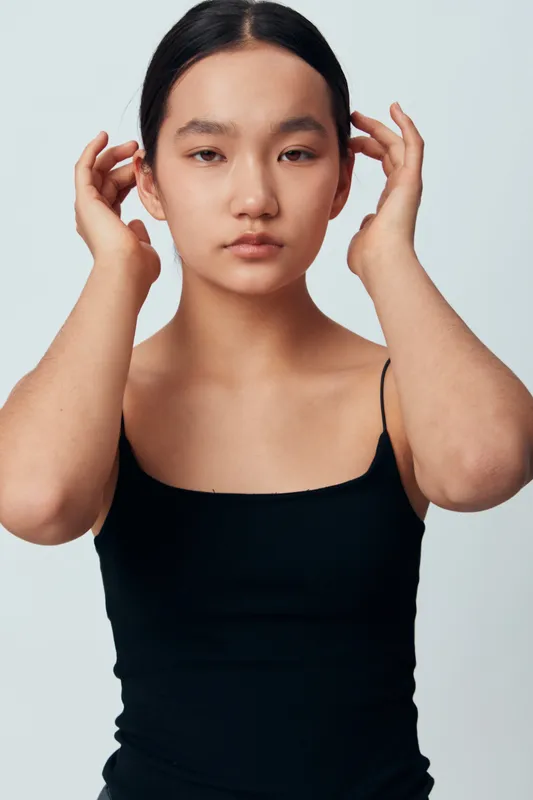 Portrait of young woman with dark hair in black top posing with hands near face on neutral background. Female model showing natural look, skin texture and minimalist style for beauty and fashion.