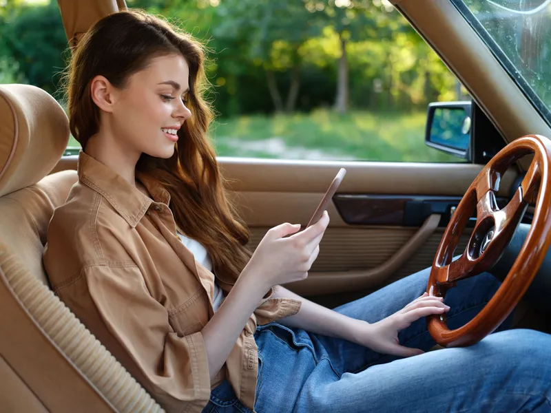 Young woman enjoying her smartphone in a vintage car, dressed casually in a beige shirt and jeans, surrounded by nature, conveying relaxation and joy.