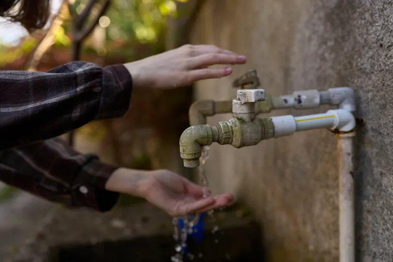 Casual woman outdoors, washing hands under a wall faucet, sunlit moment, focused gesture, clear water flow, natural colors, simple everyday scene