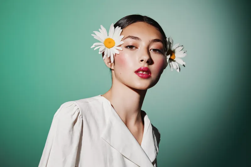 Young woman with floral ear accessories poses in a stylish white outfit against a green background. Makeup highlights her glowing skin and red shiny lips. Beauty fashion concept.