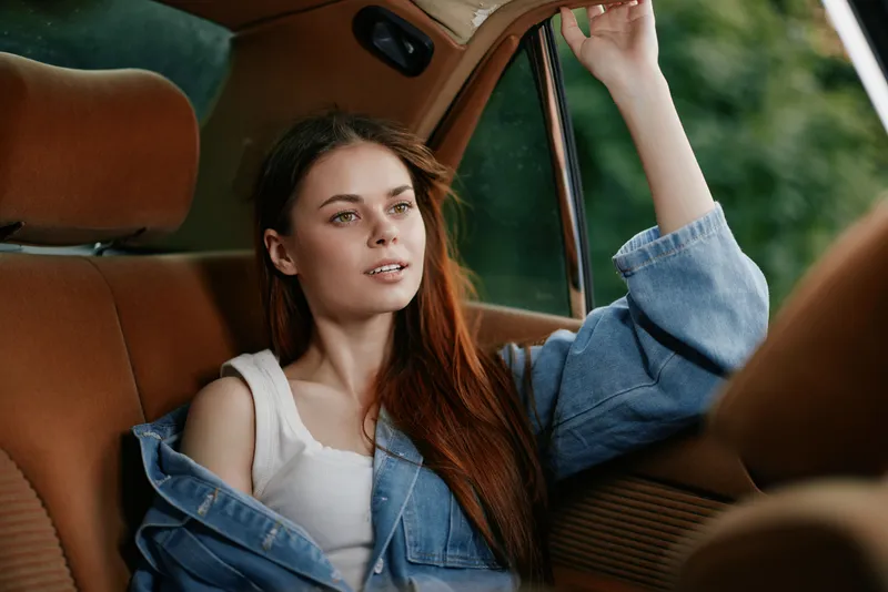 Young woman in denim jacket sitting in a car, enjoying a thoughtful moment, captured in natural light with a serene expression, reflecting a casual and relaxed lifestyle.