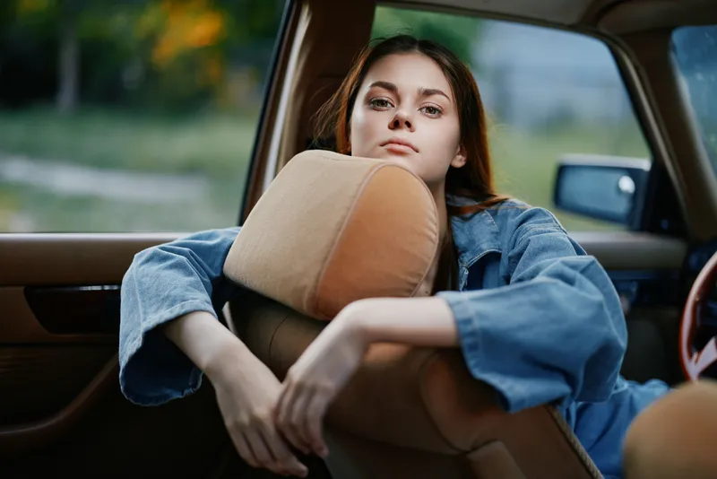 Young woman in denim jacket relaxing in a vintage car, exuding a contemplative mood with a soft, natural background highlighting vintage aesthetics.