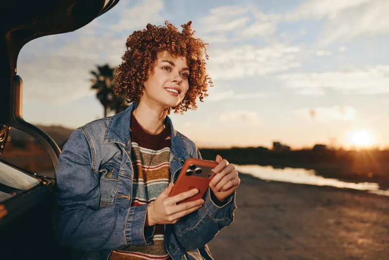 Woman lifestyle outdoors sunset smartphone denim car road, cheerful smile curly hair, leaning near vehicle door, holding phone in hands, warm golden light, soft breeze, scenic sky