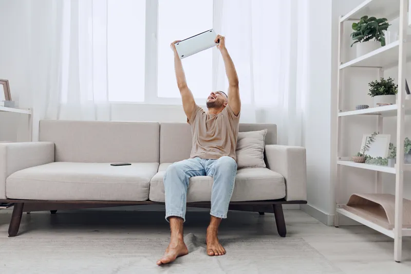 Energetic man celebrating success at home, holding a laptop high while sitting on a cozy sofa. Bright, modern interior creates an inviting atmosphere.