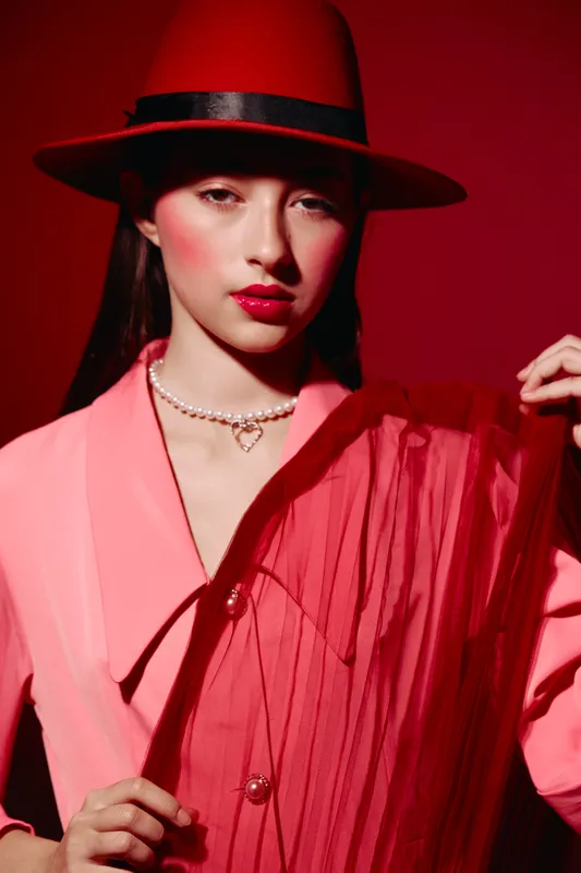 Stylish woman wearing a red hat and a pink layered outfit poses against a red background. She wears striking makeup, pearls, and exudes bold fashion and beauty creativity.