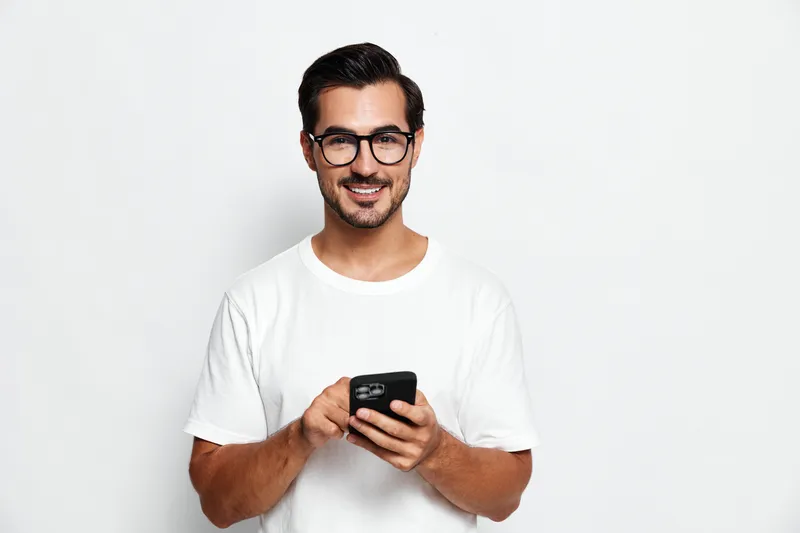 Happy smiling man wearing glasses holds smartphone in hands while looking at camera, standing against a plain solid light background. People lifestyle concept.