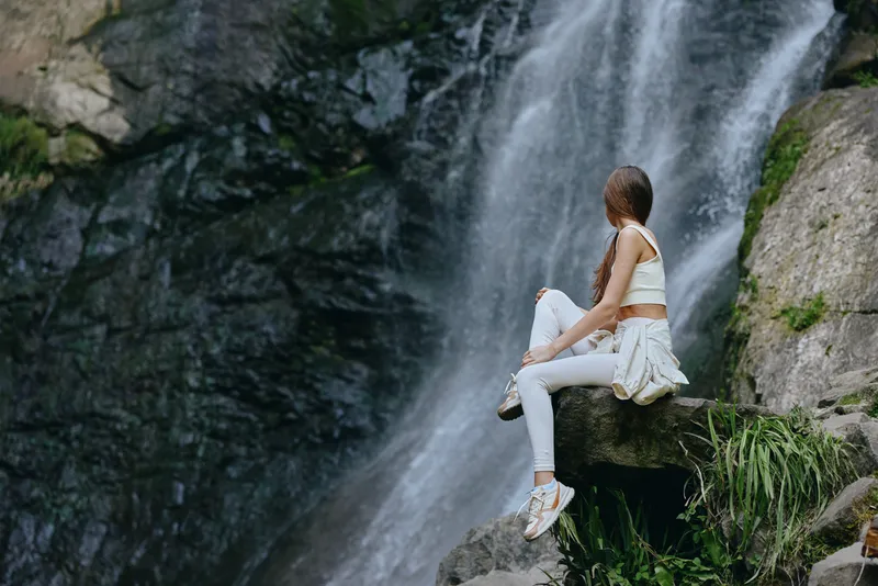 woman sitting by waterfall surrounded by rocks and greenery, wearing casual white clothes and sneakers, enjoying peaceful nature in outdoor forest environment