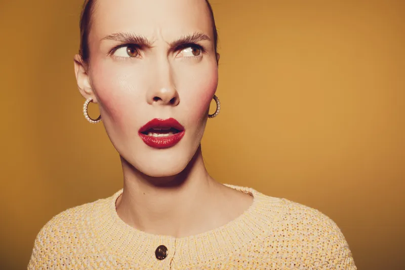 Confused woman with red lipstick and hoop earrings looks sideways with raised eyebrows. Close-up portrait on yellow background. Concept of doubt, skepticism, and uncertain emotions.
