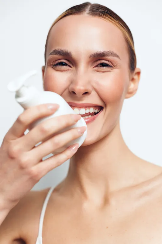 Young happy woman with clear skin smiling and holding white cosmetic bottle close to her face isolated on white background, skincare concept, beauty and health care routine.